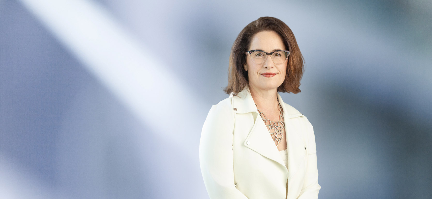 A woman, Andrea Badalucco, in a white jacket, glasses and chain link necklace is smiling at the camera, standing against a blurred blue and white background.