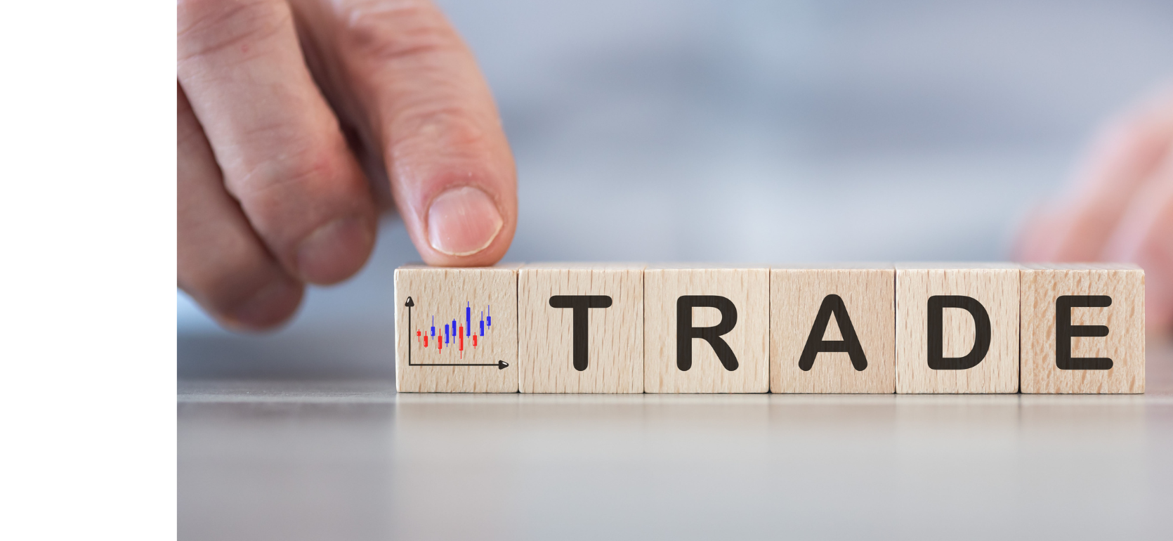 A close-up of a person’s hand placing wooden letter tiles that spell the word “TRADE” on a smooth surface. The background is blurred, focusing attention on the word.