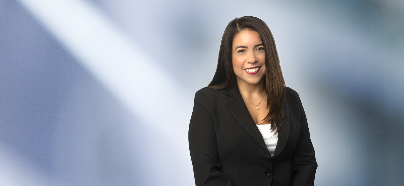An attractive woman, Jacquelyn Dupler, in a white blouse with a black blazer is smiling at the camera, standing against a blurred blue and white background.
