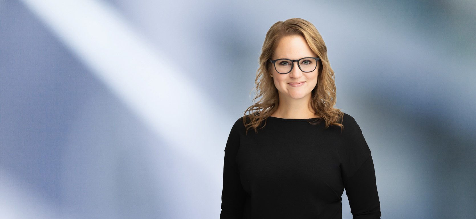 A woman, Megan Cochrane, with shoulder-length, straight blonde hair, wearing a black long-sleeved shirt and black framed glasses, is smiling at the camera, standing against a blurred blue and white background.