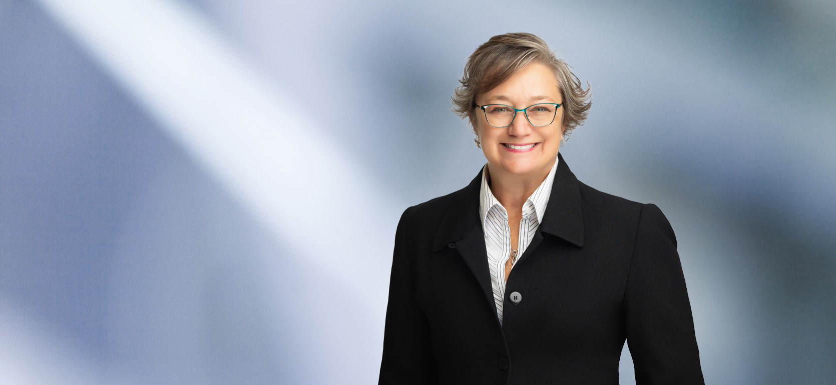 A woman, Heidi Hopper, in a black jacket with white pinstripe shirt and turquoise glasses is smiling at the camera, standing against a blurred blue and white background.
