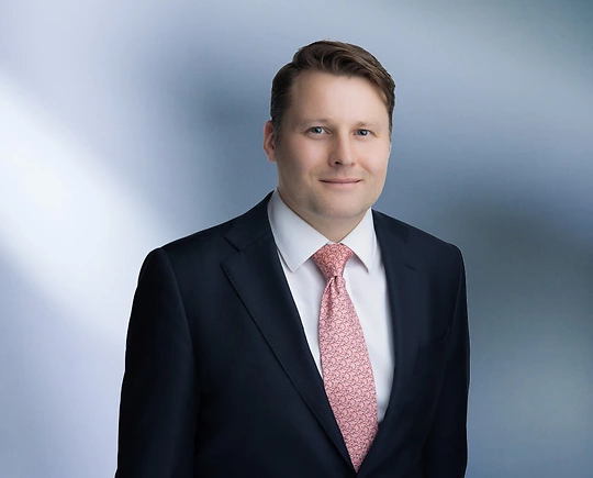 A man, Benjamin Williams, in a dark suit, white shirt, and light pink patterned tie is smiling at the camera, standing against a blurred blue and white background.