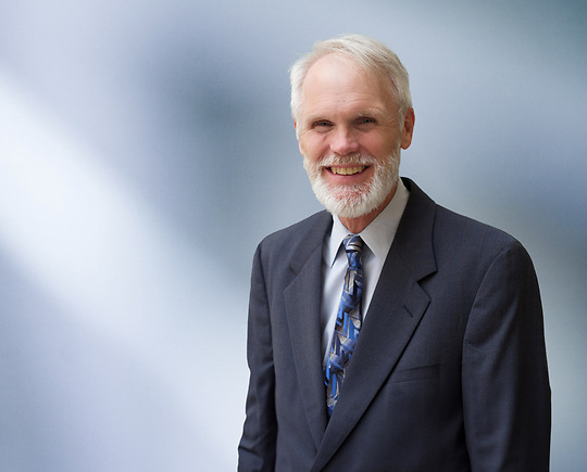 A man, Charles Barbieri, in a dark suit, white shirt, and patterned blue tie is smiling at the camera, standing against a blurred blue and white background.