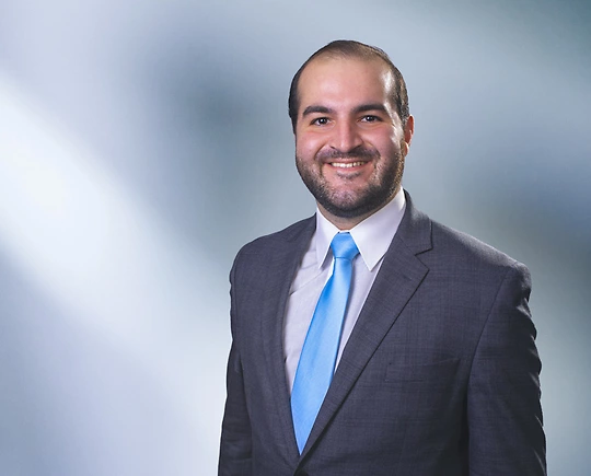 Man, Patrick Quinn, with short brown hair wearing a dark gray blazer, smiling against a blurred white and blue background.