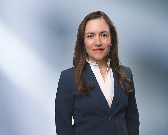 A woman, Gabrielle Lawrence, in a dark suit, light shirt, smiling at the camera, standing against a blurred blue and white background.