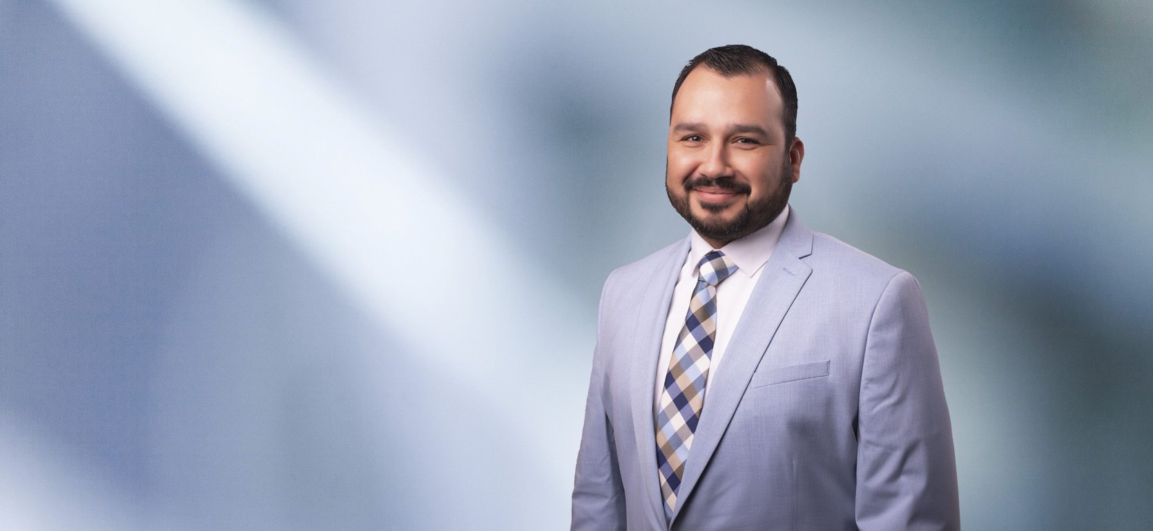 A man, Juan Negrete, in a light blue suit, white shirt, and patterned tie is smiling at the camera, standing against a blurred blue and white background.