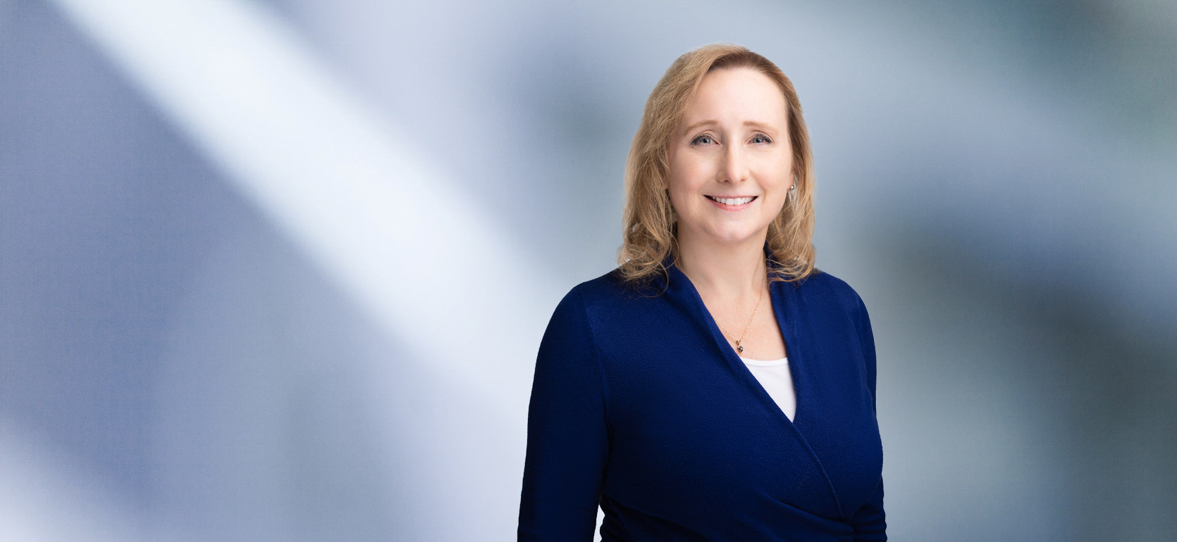 A woman, Ann Stevens, in a blue top and white blouse is smiling at the camera, standing against a blurred blue and white background.