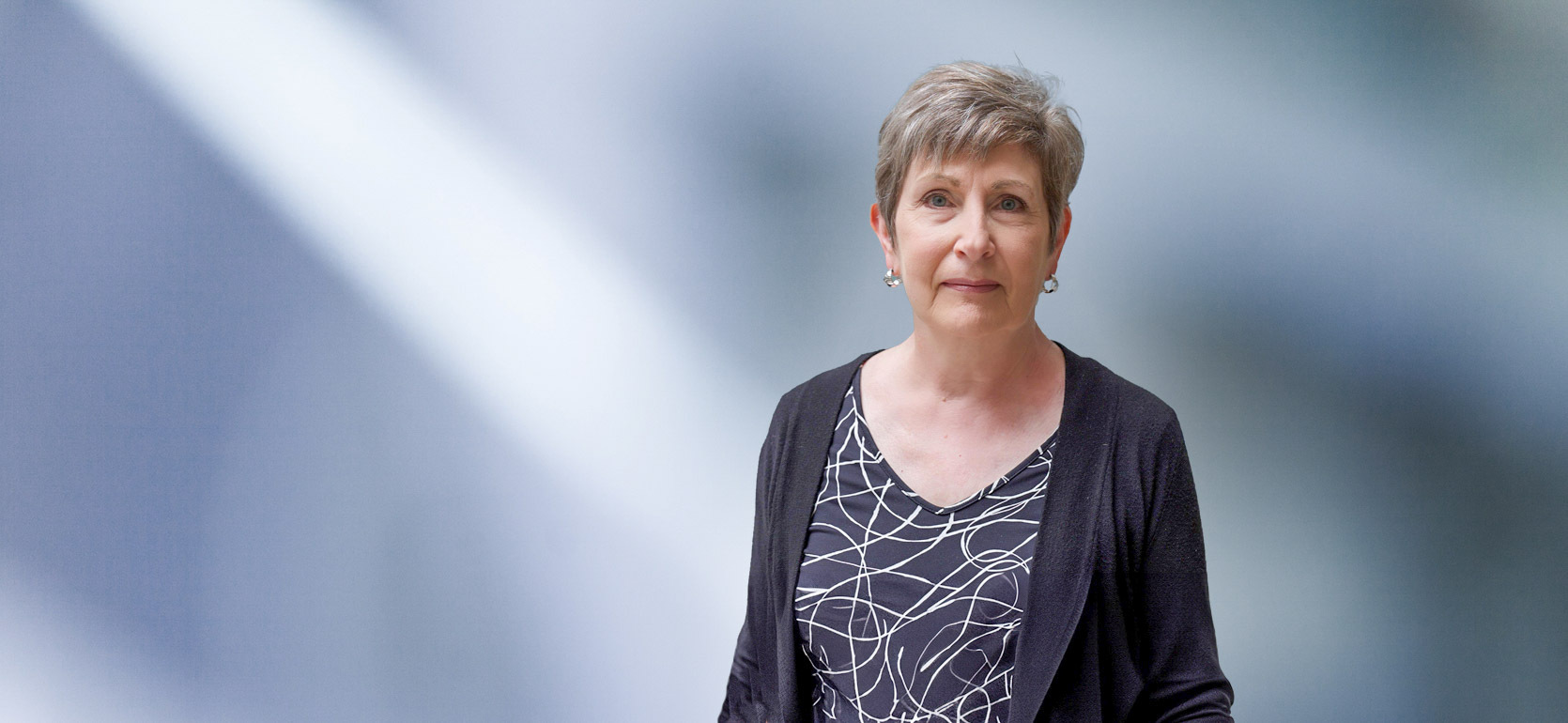 A woman, Carol Palomaki, in a black cardigan and patterned top is looking at the camera, standing against a blurred blue and white background.