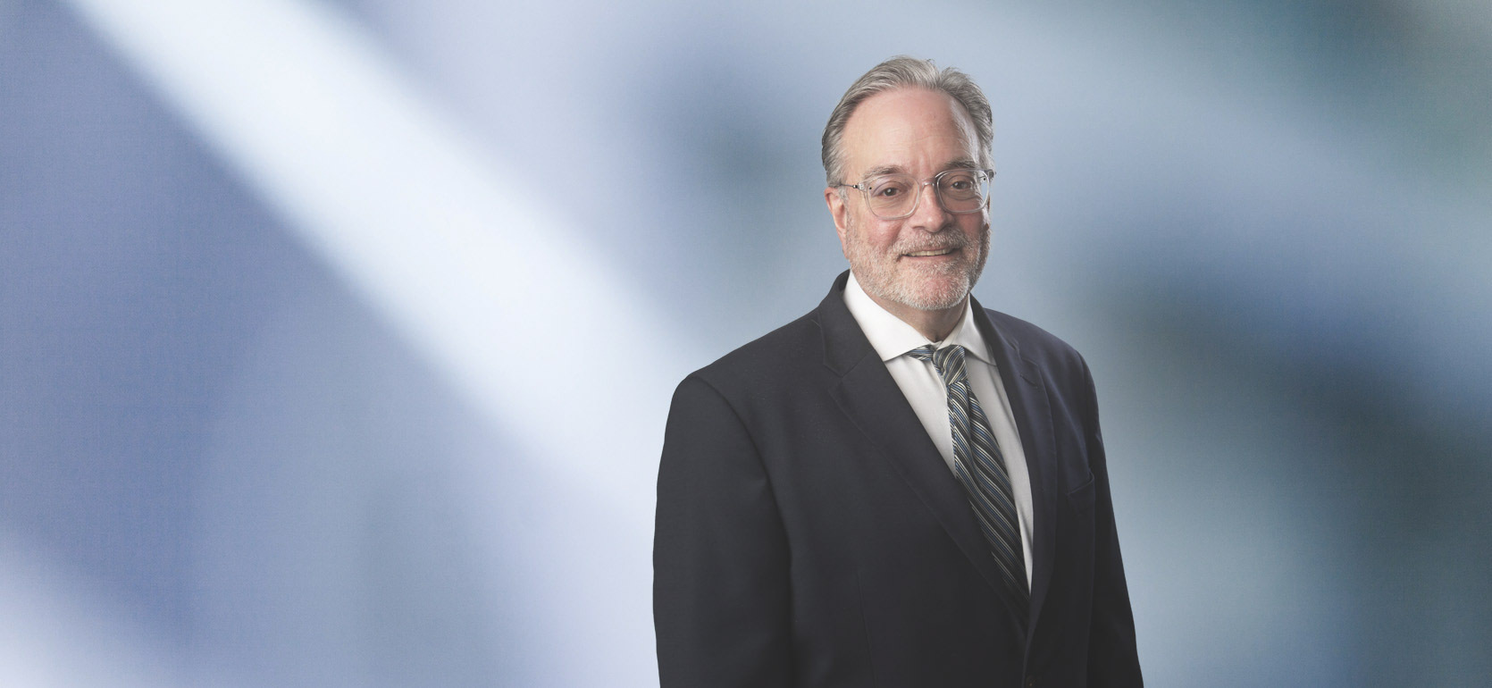 Man, Scott Chernich. wearing a dark blazer, standing against a blurred blue and white background.
