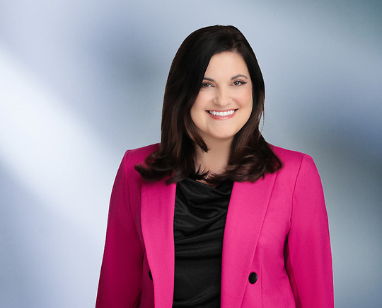 Woman, Stefania Gismondi, wearing a pink blazer, standing against a blurred blue and white background.