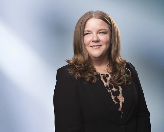 A woman, Cassandra Bowe, in a black blazer and leopard print top is smiling at the camera, standing against a blurred blue and white background.