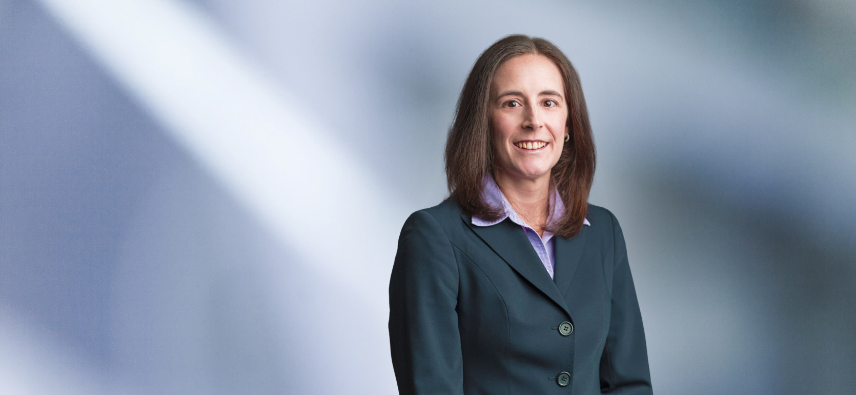 A woman, Erica Huddas, in a dark green blazer and light purple blouse is smiling at the camera, standing against a blurred blue and white background.