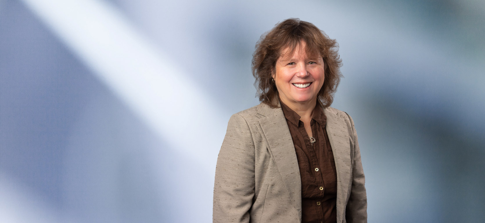 A woman, Kimberly Hafley, in a tan blazer and dark brown shirt is smiling at the camera, standing against a blurred blue and white background.