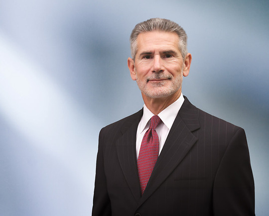 A man, Lawrence Korolewicz, in a dark pinstripe suit, white shirt, and red patterned tie is looking at the camera, standing against a blurred blue and white background.