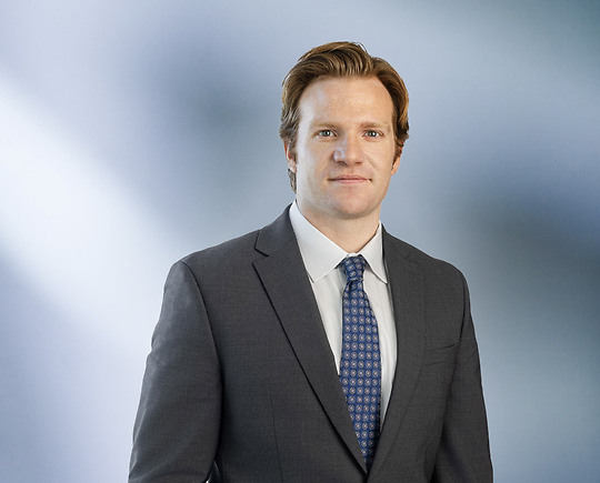 A man, Anthony Dalimonte, in a gray suit, blue shirt, and blue striped tie is smiling at the camera, standing against a blurred blue and white background.