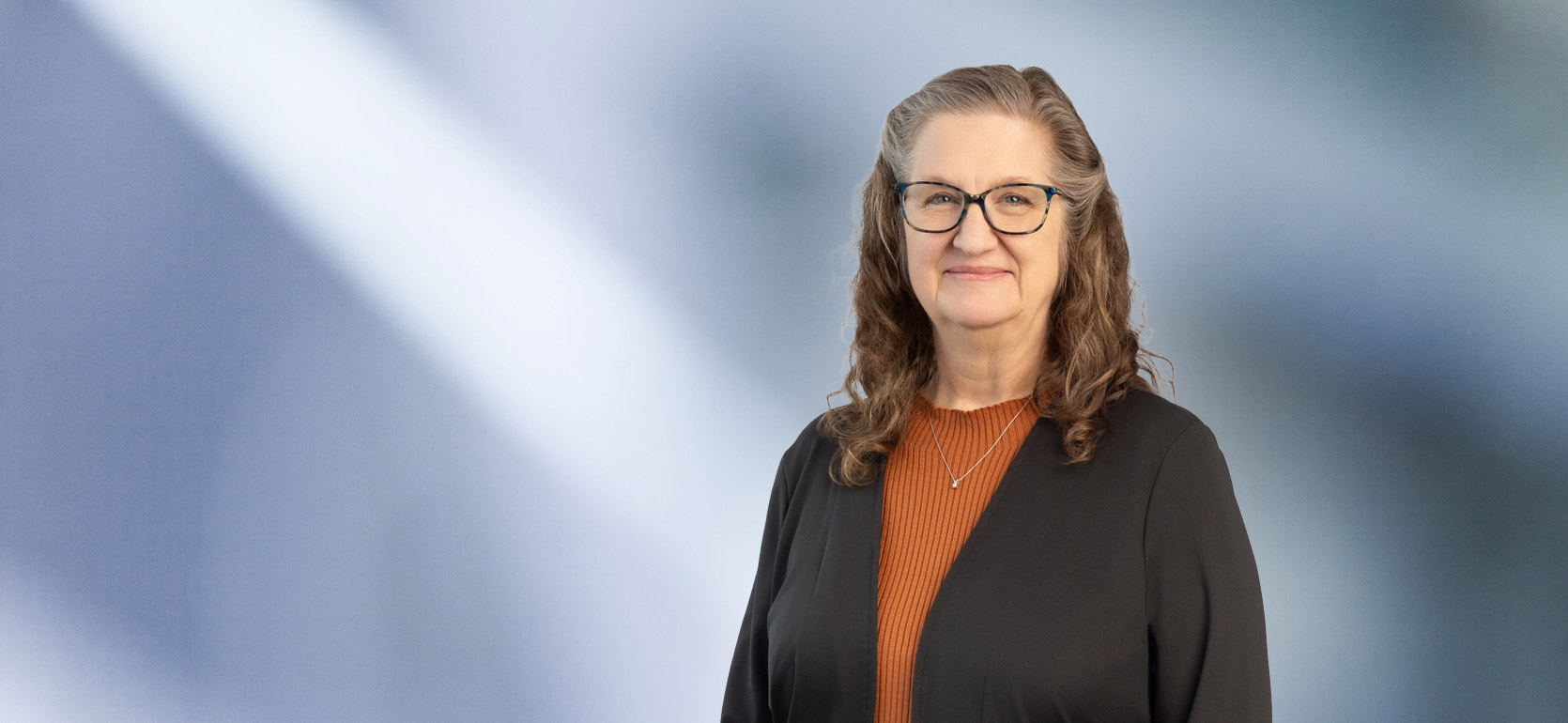 A woman, Susan Paules, in a brown sweater and dark rimmed glasses is looking at the camera, standing against a blurred blue and white background.