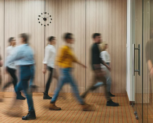 Blurred image of a group of people walking in a hallway with wooden panel walls and a modern clock, suggesting motion, transition, or a busy office environment.