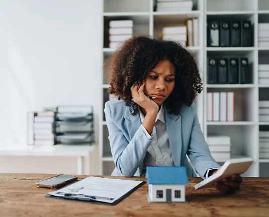 Woman looking at a calculator with concern. A phone, clipboard, and a model house sit on the table before her.
