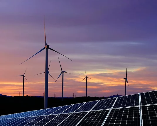 Landscape at sunset featuring several wind turbines and rows of solar panels. The sky displays a gradient of purple, orange, and blue hues.