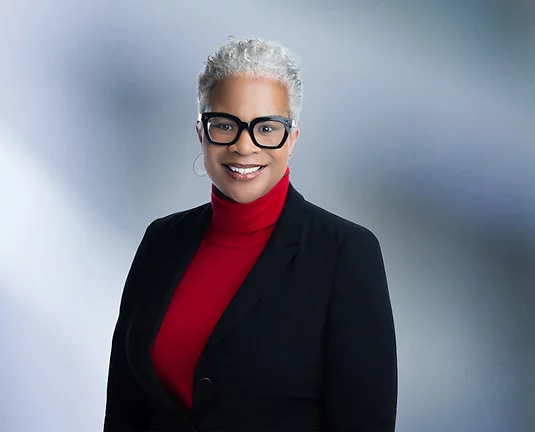Older African American women wearing black blazer with red turtleneck with thick dark rimmed glasses smiles at camera