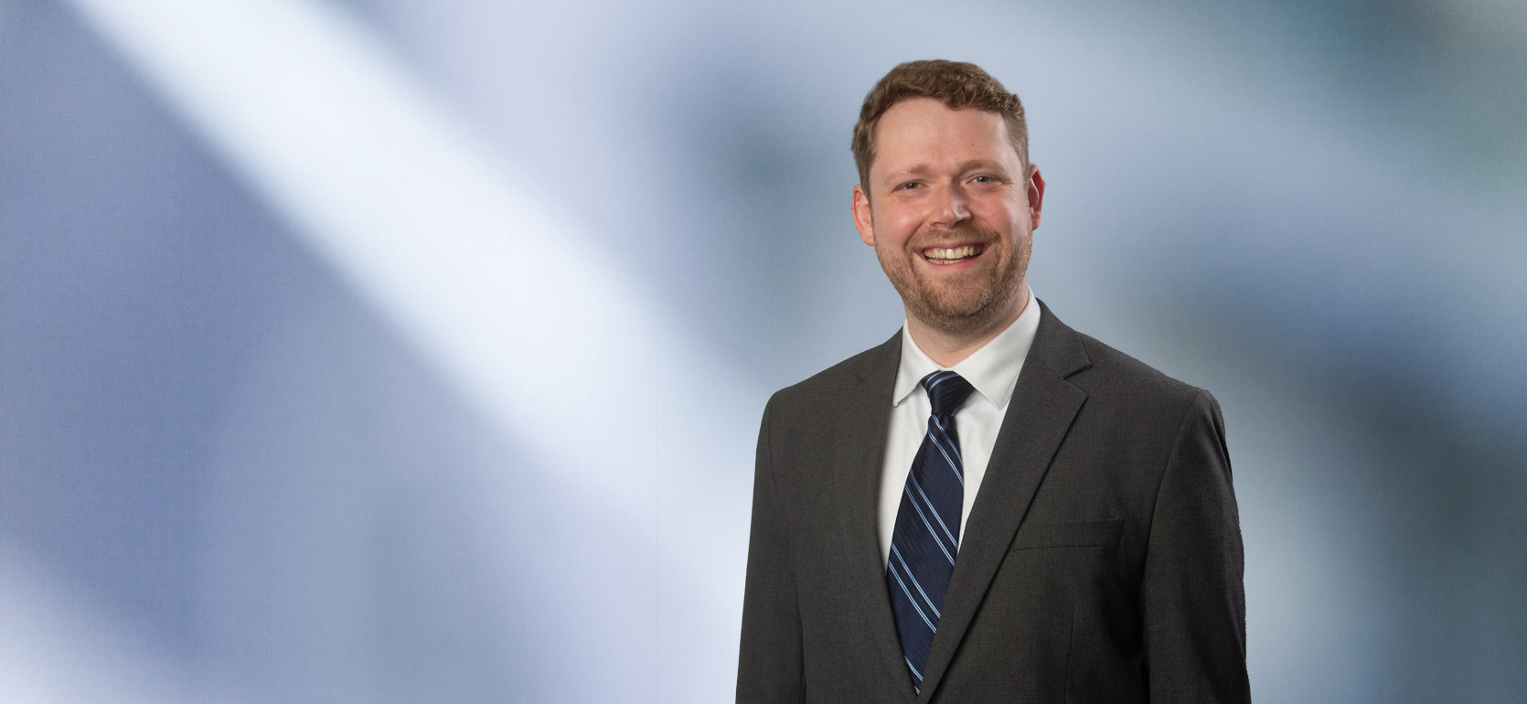 Man with beard, Jon Austin, smiles at camera wearing a gray sport coat and grey striped tie in front of blue gray background