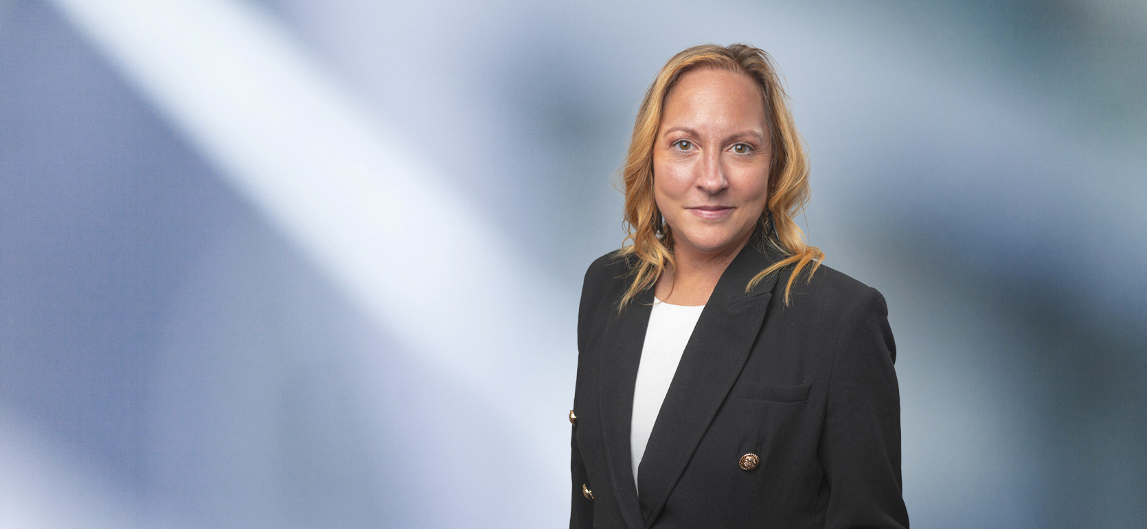 A woman, Elizabeth Cotner, in a black blazer with gold buttons and a white top is looking at the camera, standing against a blurred blue and white background.