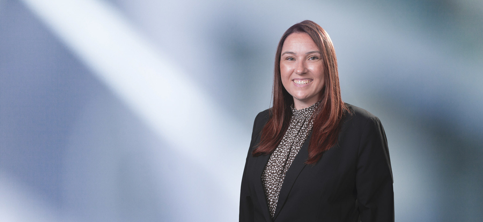 Woman, Stephanie Frizzell, wearing a dark blazer, standing against a blurred blue and white background.