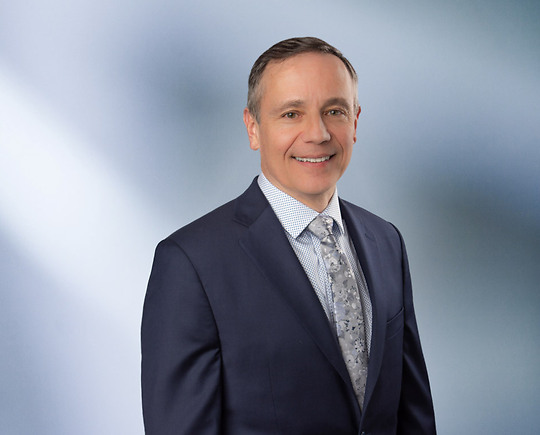 “A man, Matt Hrebec, with short, light brown hair, wearing a grey blazer and white collared shirt, is smiling at the camera, standing against a blurred blue and white background.”