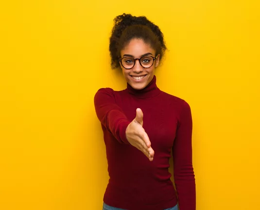 Confident young woman wearing glasses and a red turtleneck offering a handshake while smiling, standing against a bright yellow background.