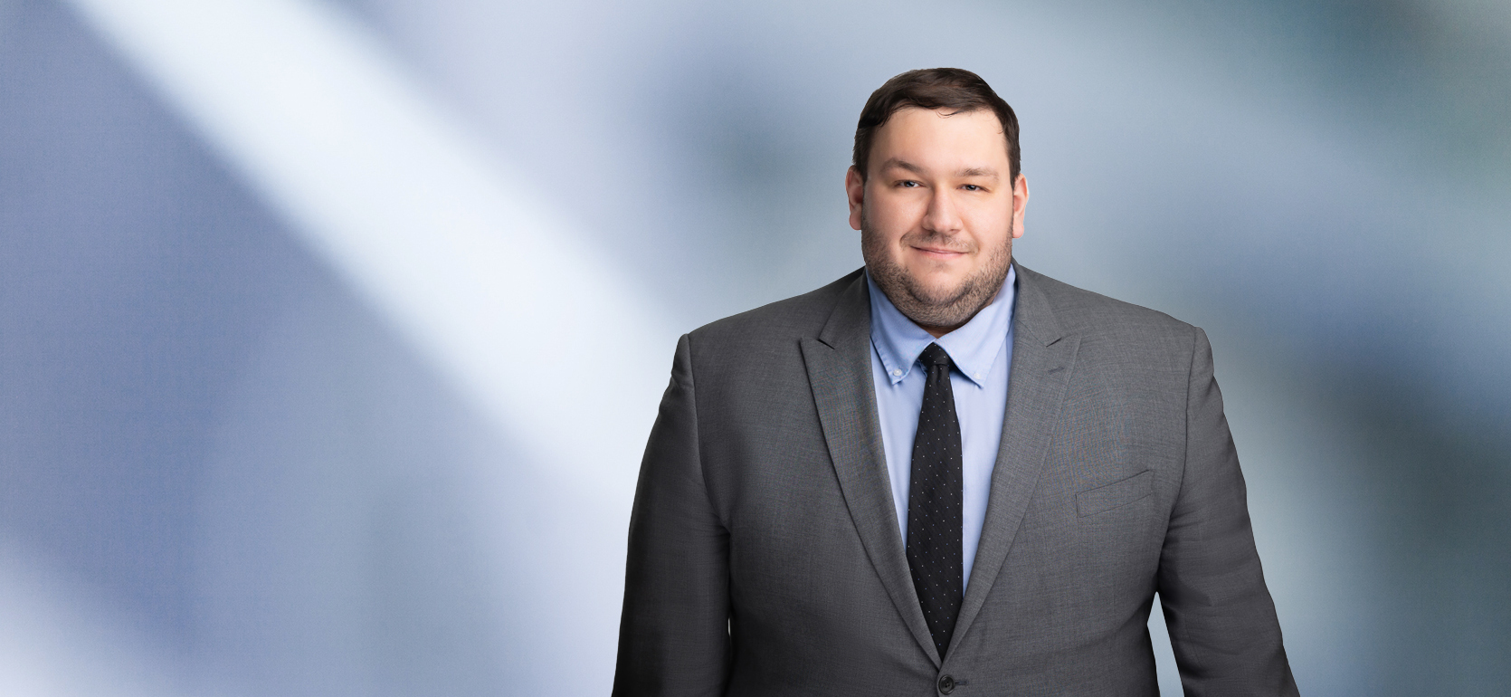 A man, Brandon Schumacher, in a gray suit, light blue shirt, and black tie is smiling at the camera, standing against a blurred blue and white background.