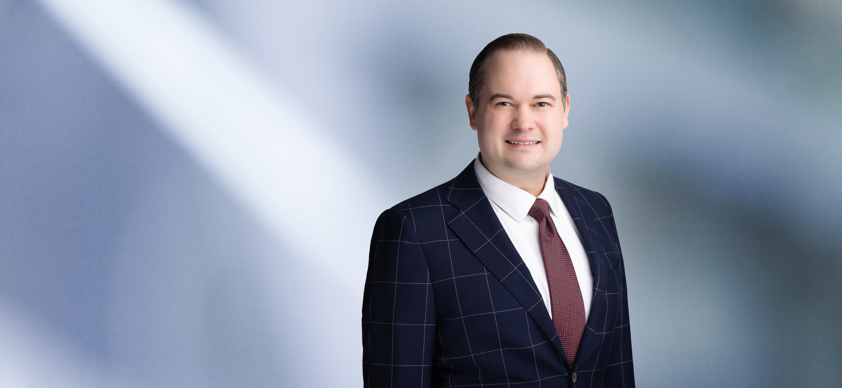 A man, Alexander Rusek, in a dark suit, light shirt, and patterned tie is smiling at the camera, standing against a blurred blue and white background.