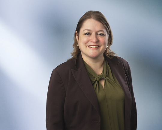 A woman, Anna Gibson, in a dark blazer and green blouse is smiling at the camera, standing against a blurred blue and white background.