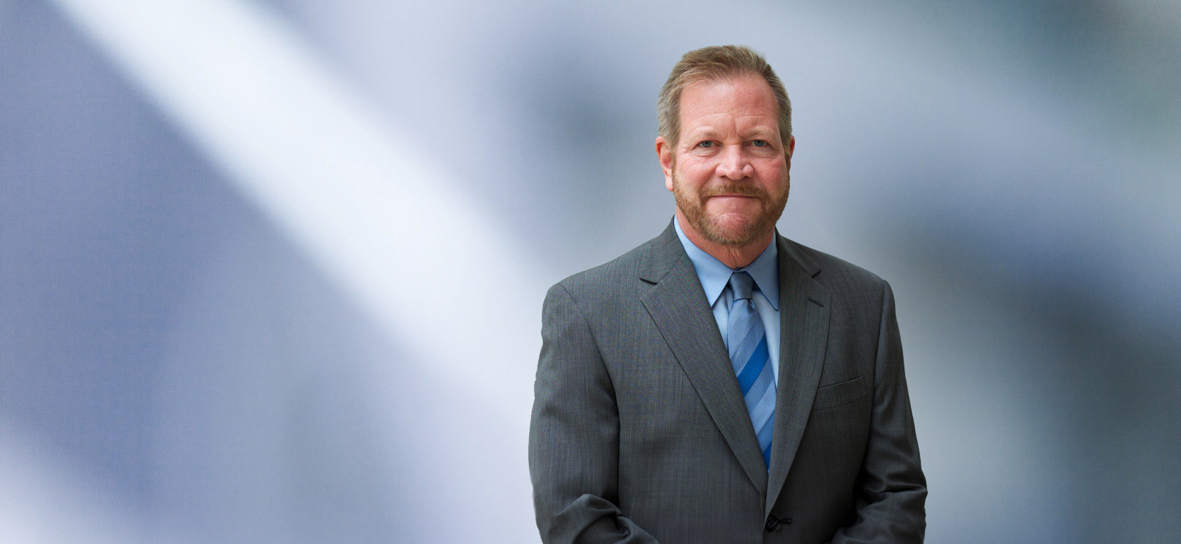 A man, Charles Janssen, in a gray suit, light blue shirt, and blue striped tie is looking at the camera, standing against a blurred blue and white background.