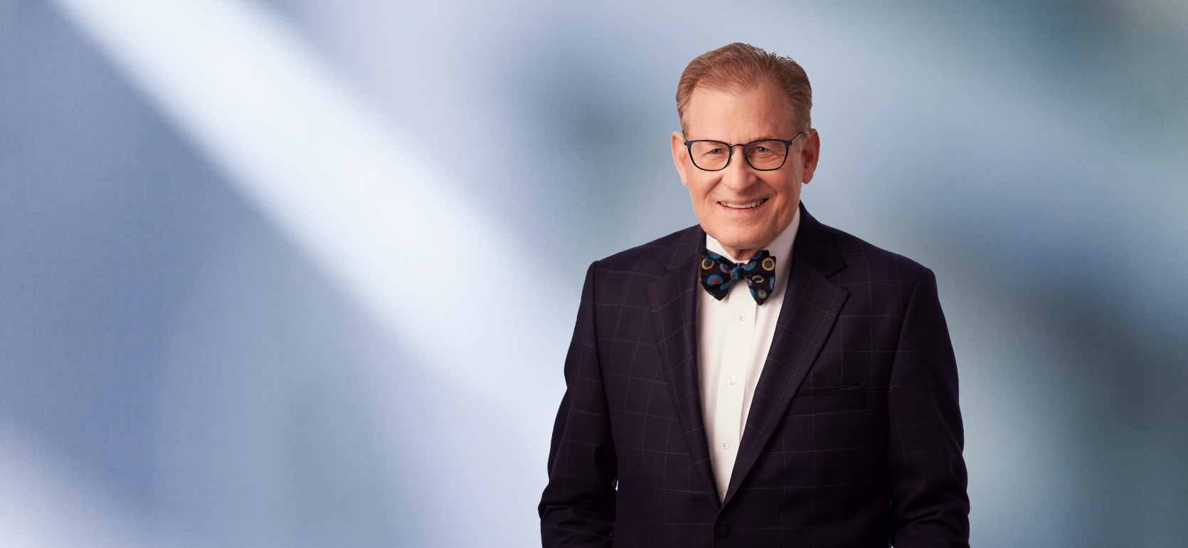 A man, Fredrick Dilley, in a dark checkered suit and a colorful bow tie is smiling at the camera, standing against a blurred blue and white background.