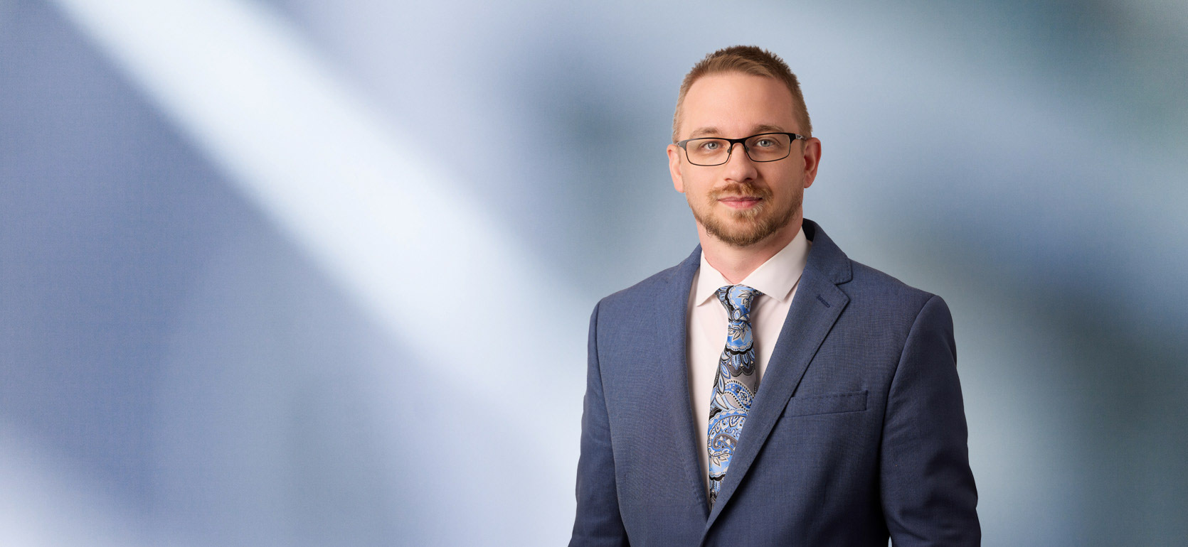 A man, Keith Brown, in a blue suit, white shirt, and patterned blue tie is looking at the camera, standing against a blurred blue and white background.