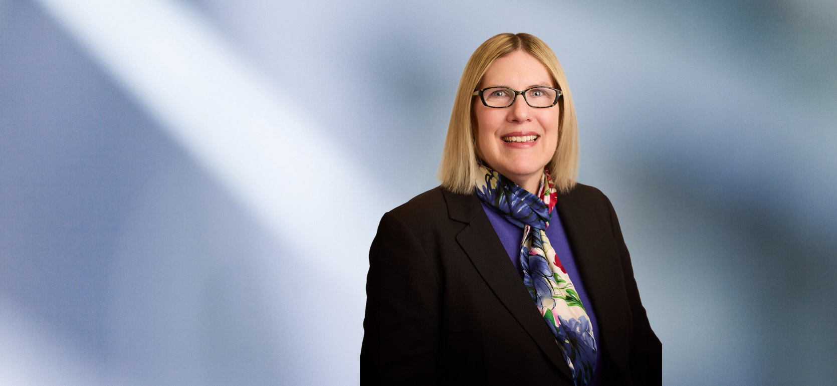 A woman, Jennifer Vanregemorter, in a black blazer, purple top, and multicolored floral scarf is smiling at the camera, standing against a blurred blue and white background.
