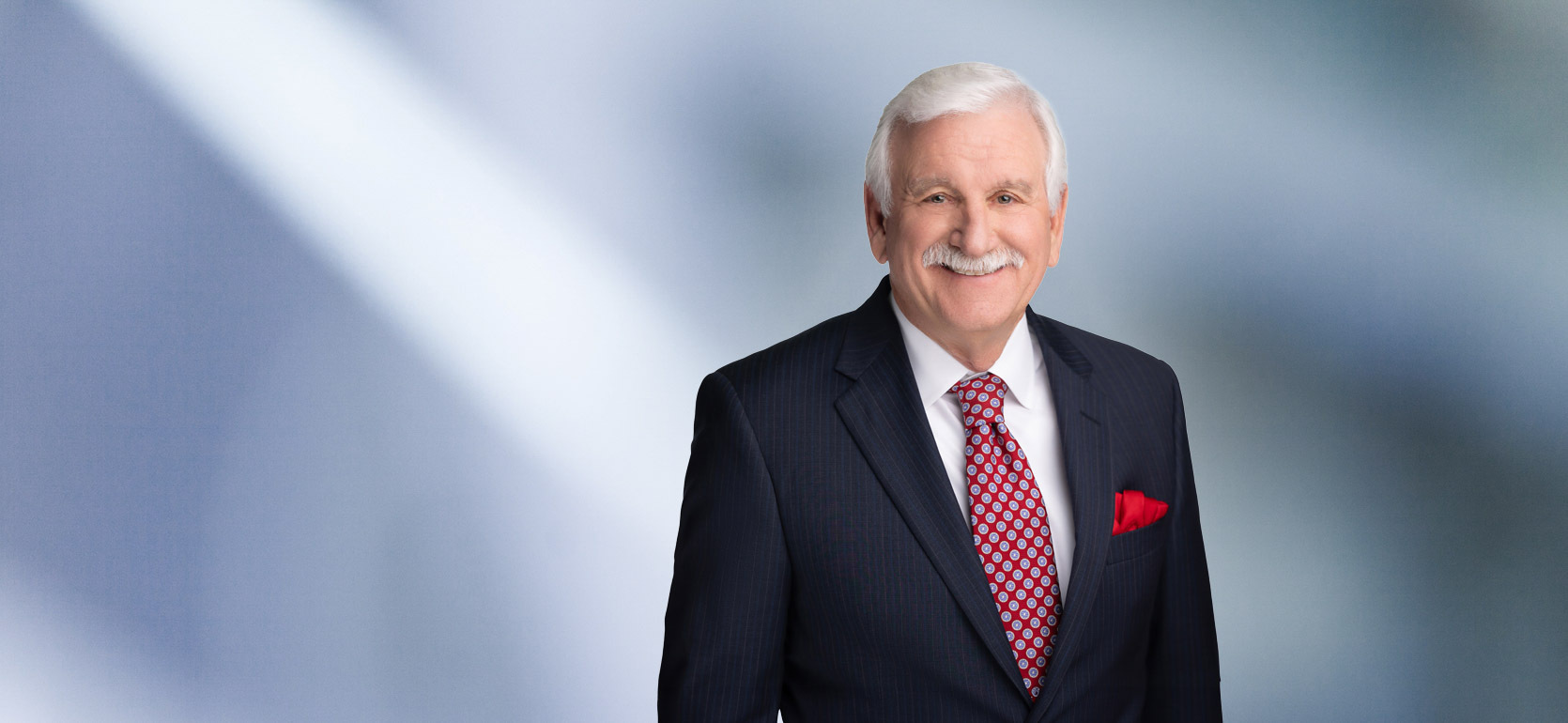 A man, Michael Kluck, with short, graying hair, wearing a dark suit and striped tie, is smiling at the camera, standing against a blurred blue and white background."