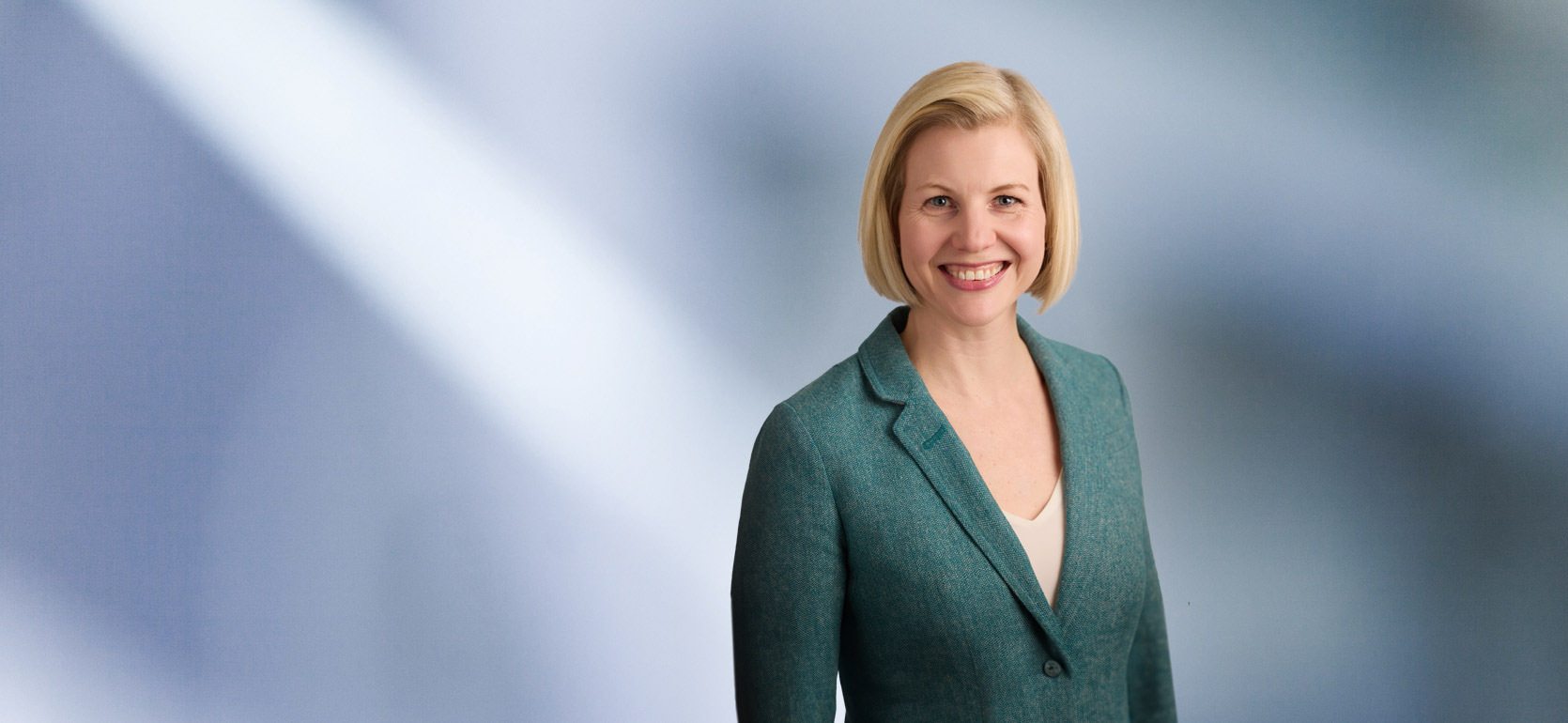 Woman, Mindi Johnson, with shoulder-length blonde hair wearing a black blazer, smiling against a blurred white and blue background.