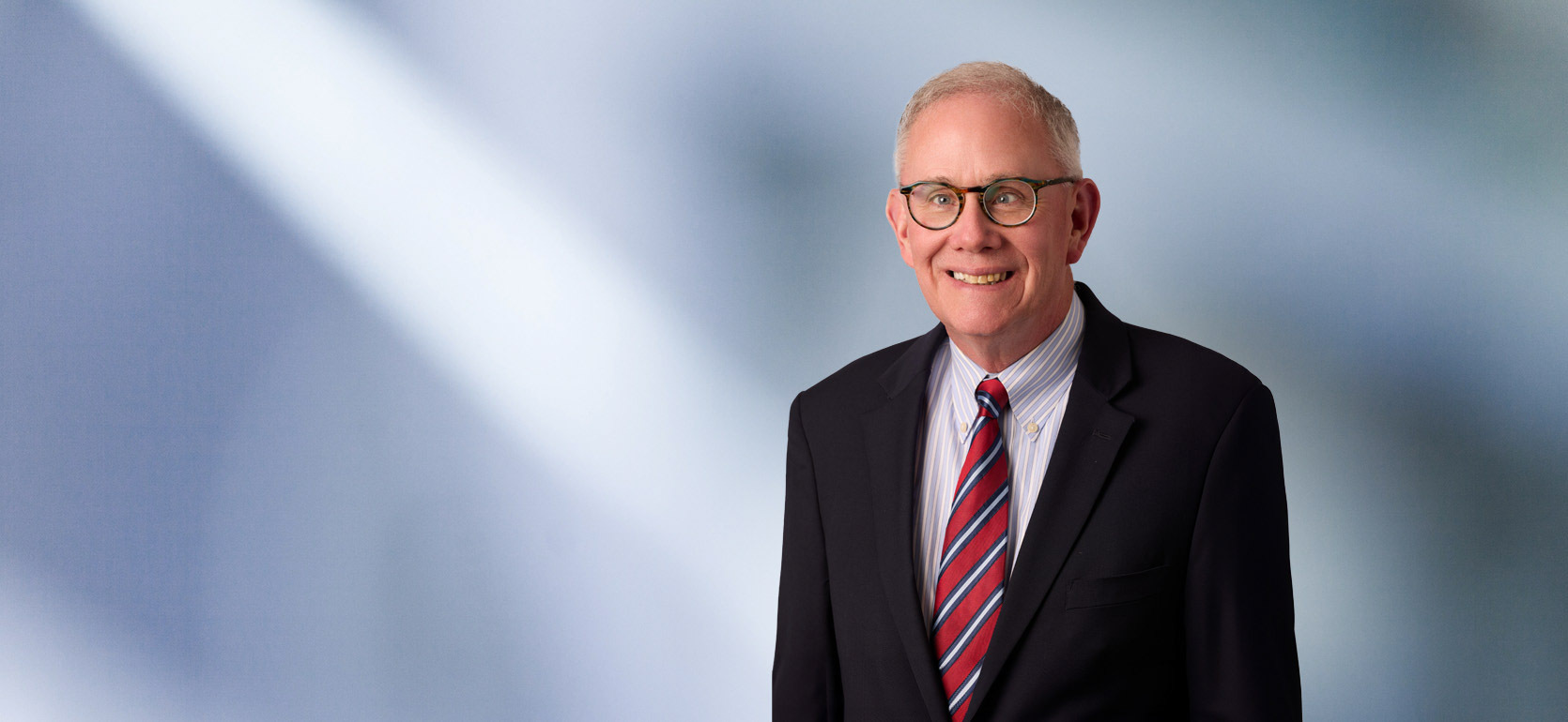 Man, Scott Hogan, wearing a dark blazer, standing against a blurred blue and white background.