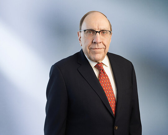man, Brian Renaud, wearing a dark suit, white shirt, red patterned tie, and glasses, standing against a softly blurred blue and white background, looking confidently at the camera