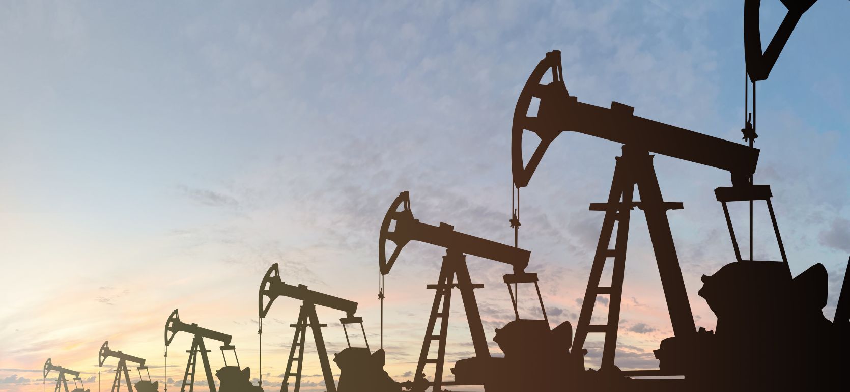 Silhouettes of multiple oil pumpjacks operating in a row against a colorful evening or early morning sky, with soft clouds and a gradient of blue, pink, and orange hues