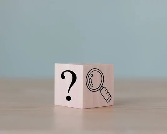 Wooden cube on a light wooden surface with a question mark symbol on one side and a magnifying glass icon on the adjacent side, against a plain light blue background.