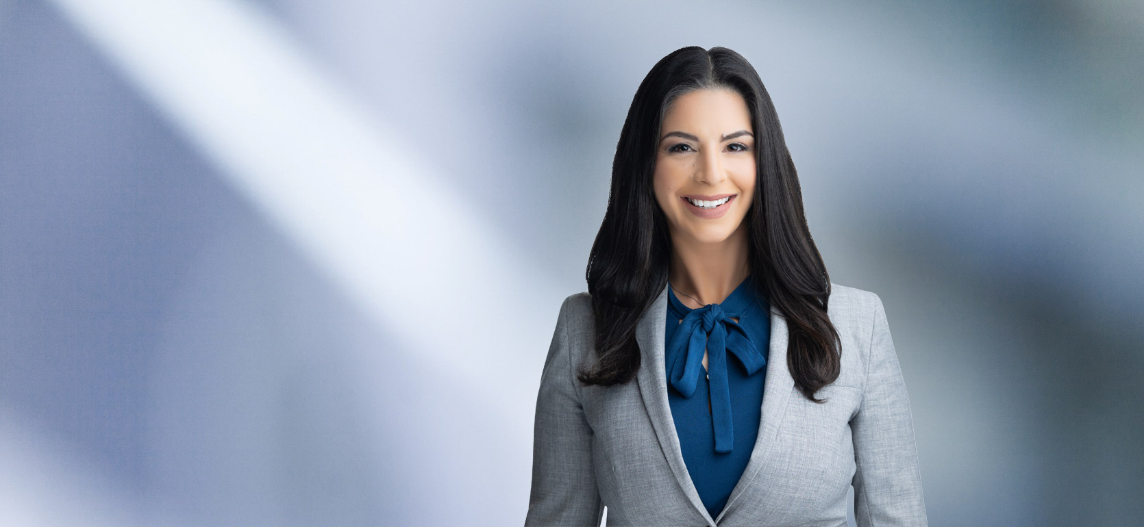A woman, Courtney Agrusa, in a light gray blazer and blue blouse with a bow is smiling at the camera, standing against a blurred blue and white background.
