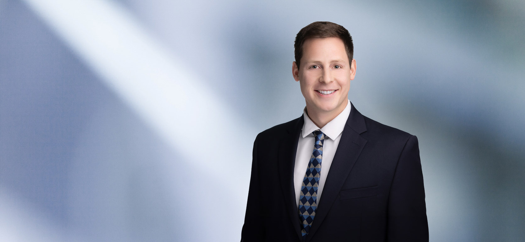 A man, Michael Cassar, with short, dark brown hair and a beard, wearing a black blazer and white shirt, is smiling at the camera, standing against a blurred blue and white background.”