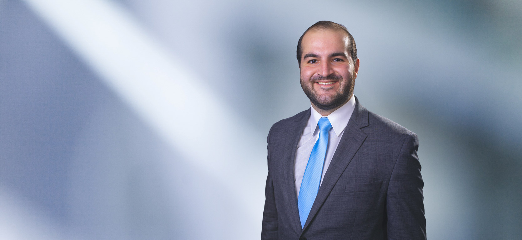 Man, Patrick Quinn, with short brown hair wearing a dark gray blazer, smiling against a blurred white and blue background.