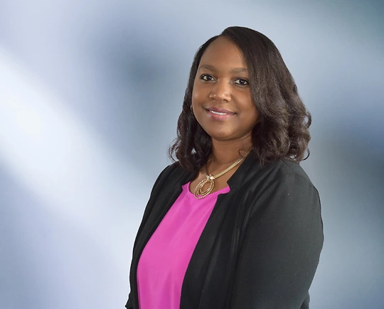 woman, Rekela Wilson, with dark brown hair, wearing a black blazer, smiling against a blurred blue and white background"