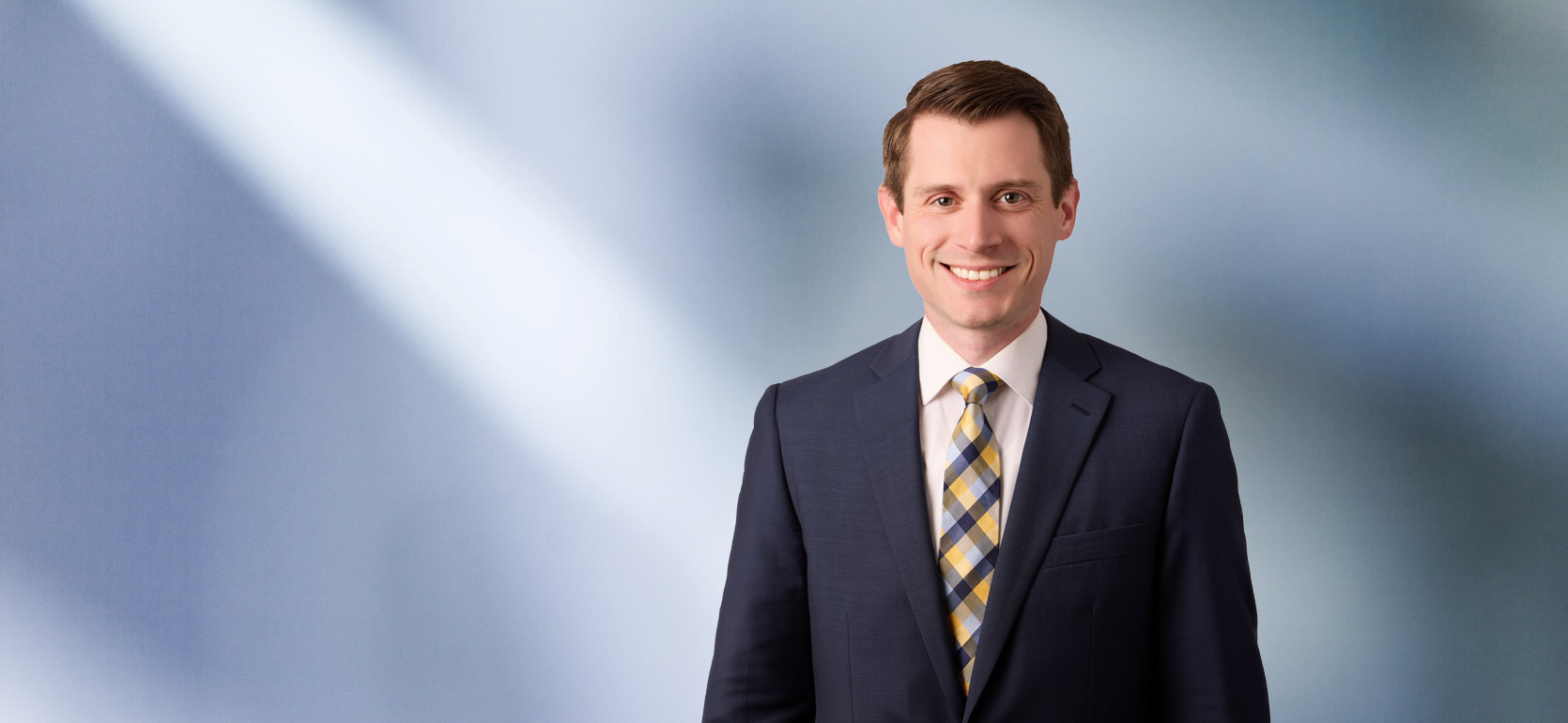 Portrait of a man, Thomas Dillon, with short brown hair in a dark blazer, with a blurred blue and white background.