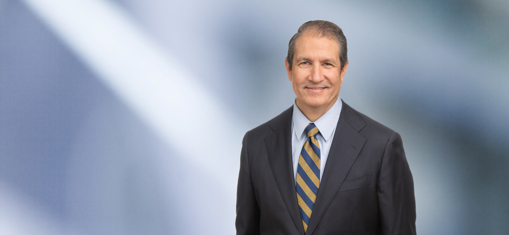 A man, Karl Butterer, in a dark checkered suit, white shirt, and blue striped tie is looking at the camera, standing against a blurred blue and white background.
