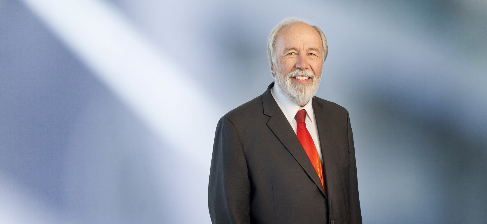 man, Dirk Beckwith, with a white beard wearing a dark suit, white shirt, and bright red tie, standing against a softly blurred blue and white background, smiling warmly at the camera.
