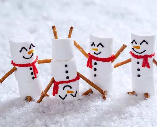 Four cute snowmen made of marshmallows with pretzel stick arms, red scarf decorations, and happy drawn-on faces, standing on artificial snow. One snowman is upside down, playfully posed.